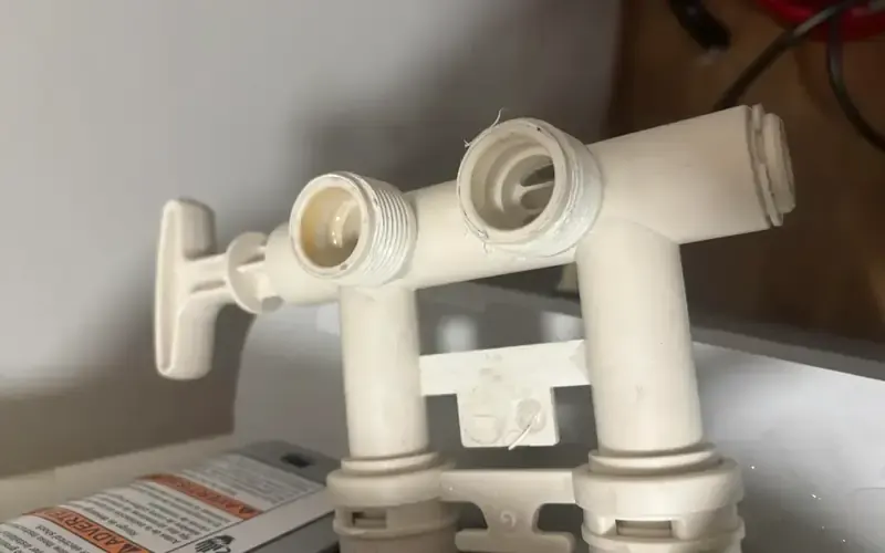 Removed water softener bypass valve close-up — white plastic body with stuck shut-off lever, cracked threading visible at inlet and outlet ports